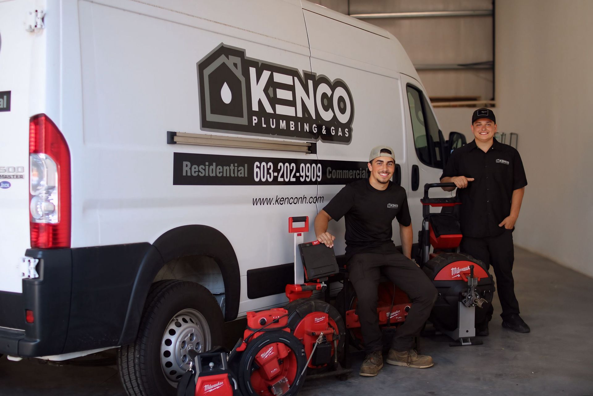 Two men pose beside a Kenco Plumbing van in a garage. They smile, with red equipment nearby.