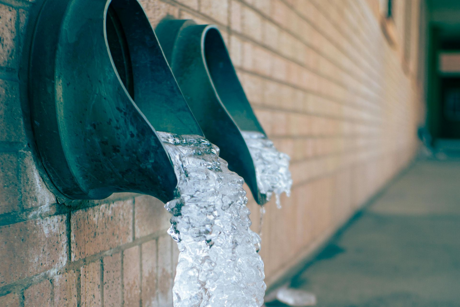 Frozen water pouring from metallic pipes on a brick wall.