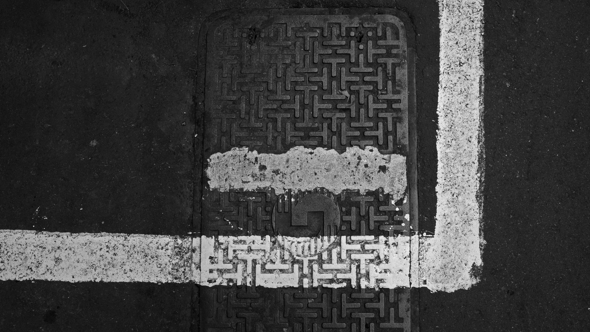 A black and white overhead view of a manhole cover and white road markings on asphalt.