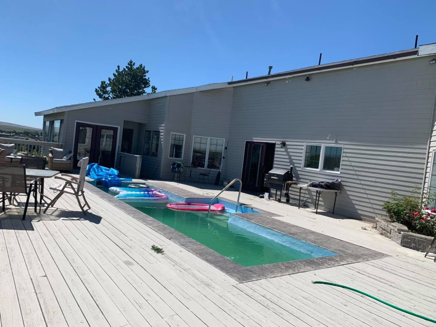 Backyard with a rectangular pool, surrounded by a wood deck, next to a house under a clear, blue sky.