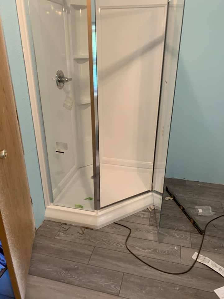 White corner shower stall with a partially open glass door, on gray tile flooring, next to a pale blue wall.