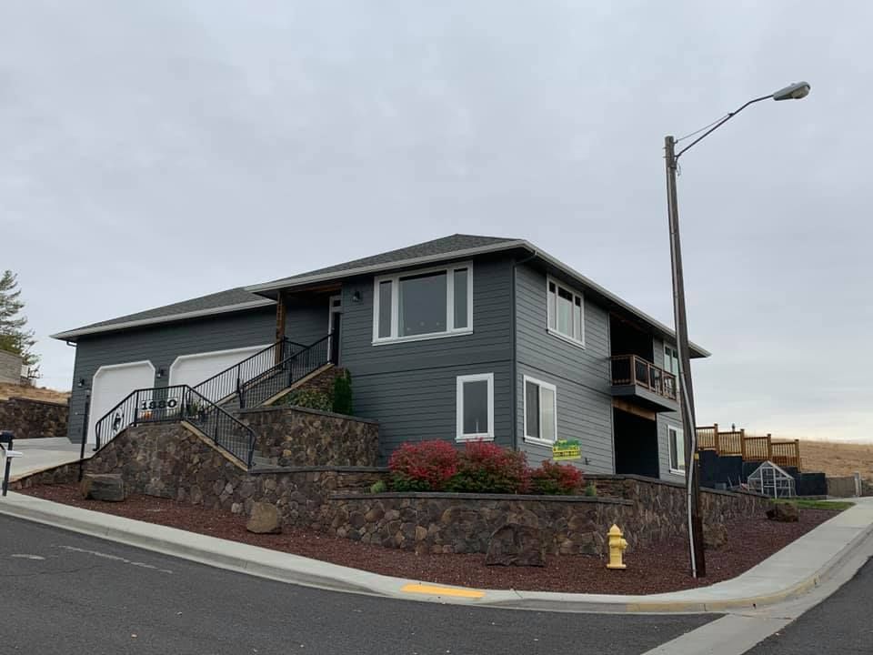 Two-story dark gray house with white trim, a stone retaining wall, and a street lamp on a cloudy day.