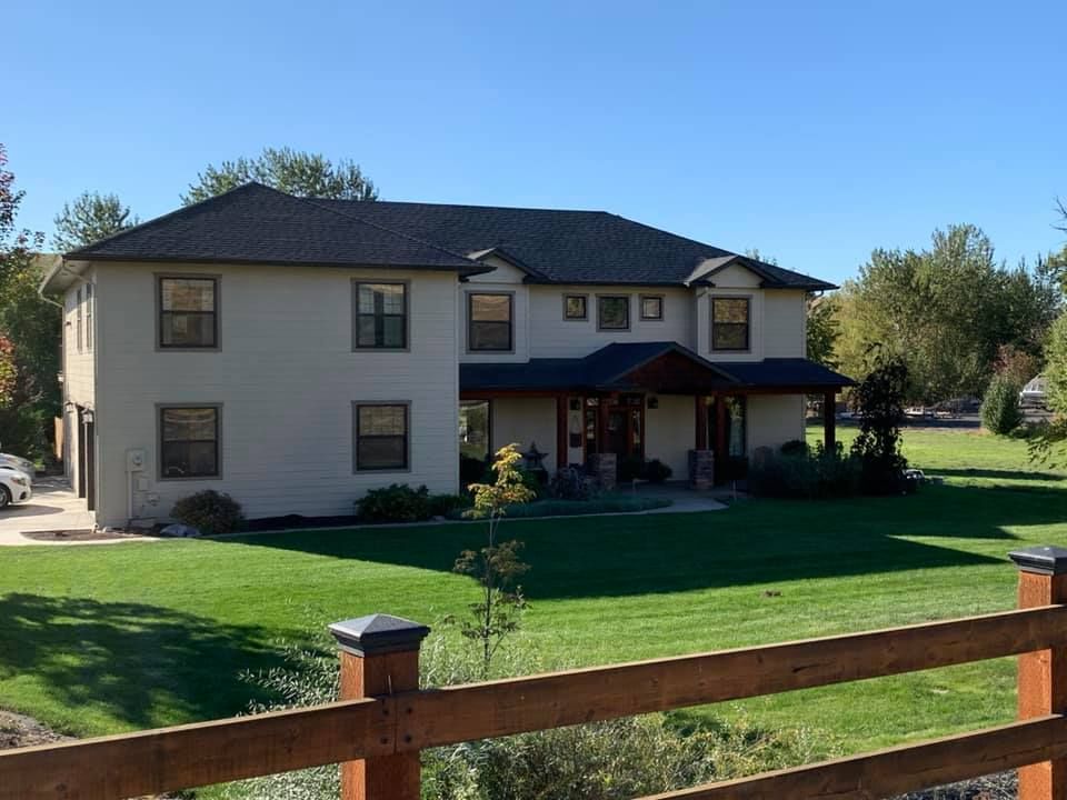 Two-story beige house with dark roof and brown trim, on a green lawn with a wooden fence.
