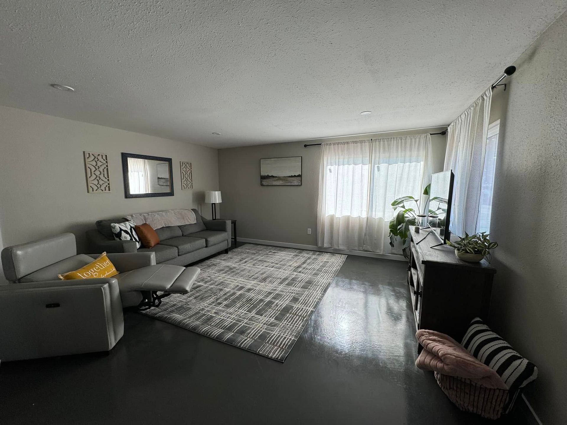 Living room with gray couch, rug, and dark floor. Curtained window, decor, and TV stand present.