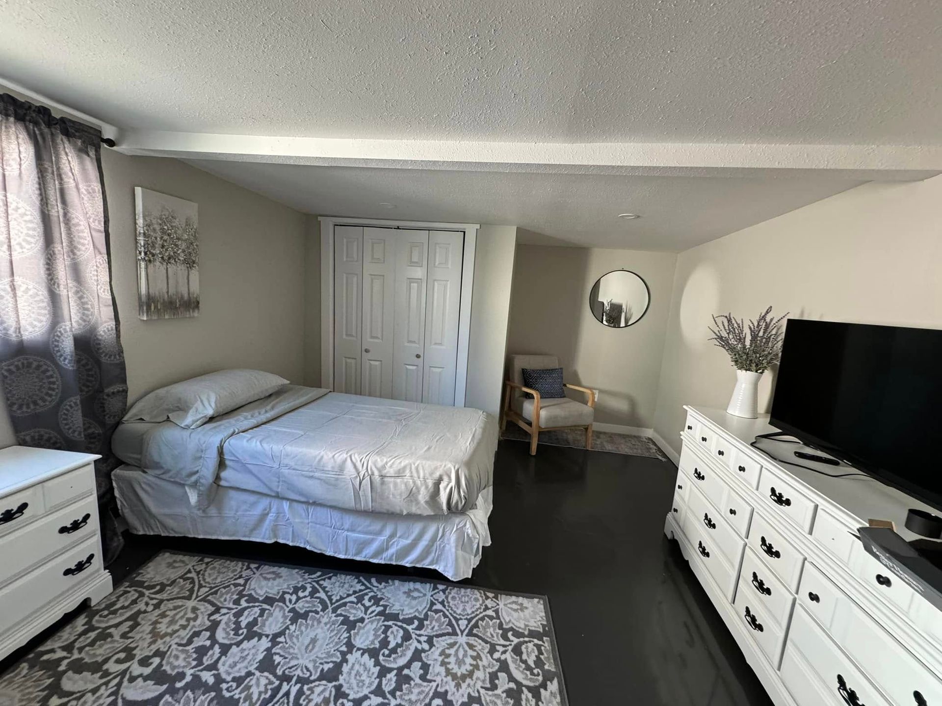Bedroom with bed, white dresser, dark floor, closet, and a chair.