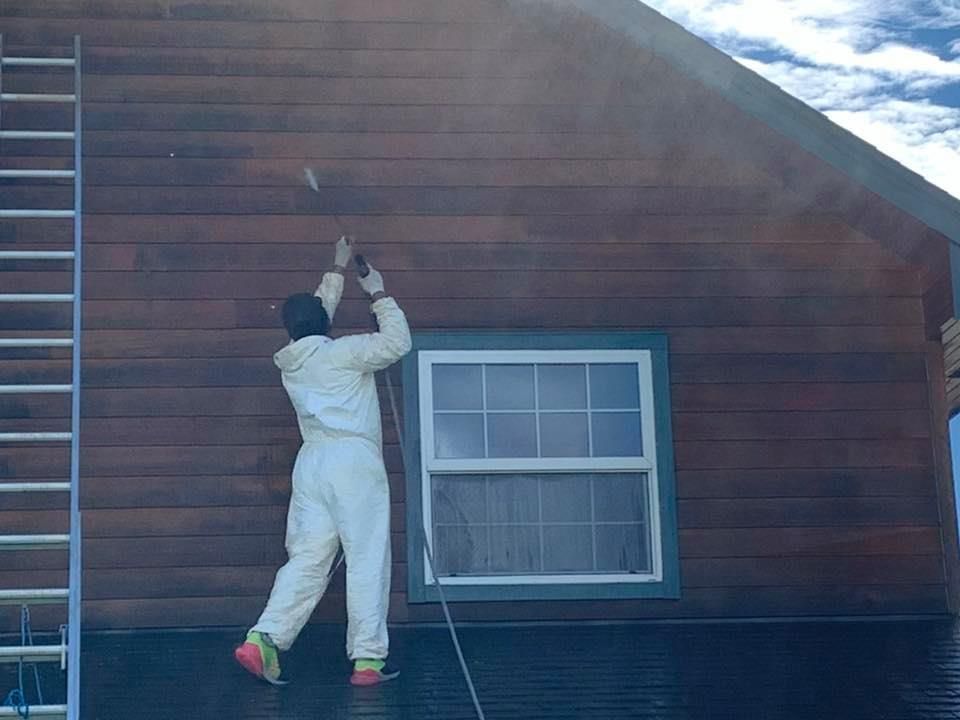 Person in protective suit power washing a dark wood exterior of a house near a window; ladder on the left.