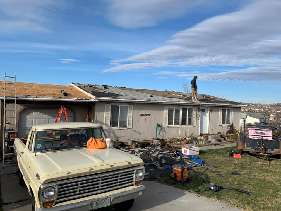 Workers re-roofing a light-colored, single-story house. An older beige pickup truck sits in the driveway. Clear sky.