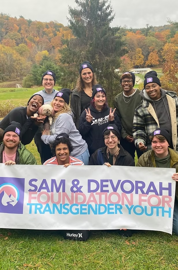 Group of people outdoors holding a banner for a transgender youth foundation, smiling. Autumn trees in the background.