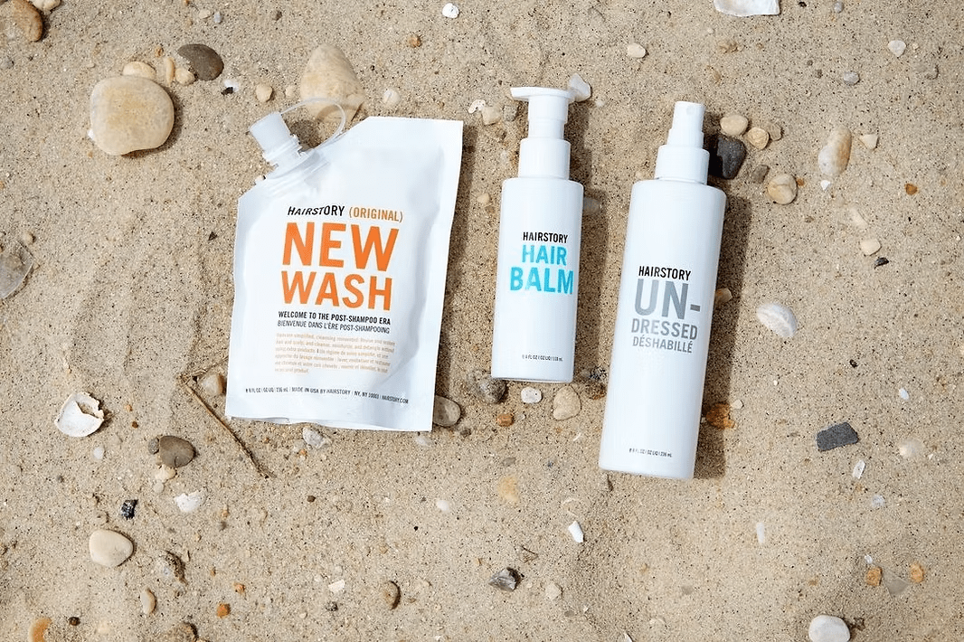 White hair care products on sand: refill pouch, hair balm, and spray.