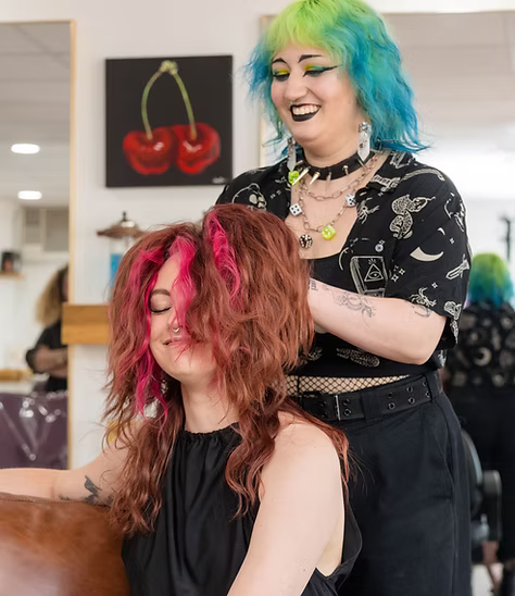 Hair stylist with blue and green hair styles a customer’s red and pink hair in a salon.