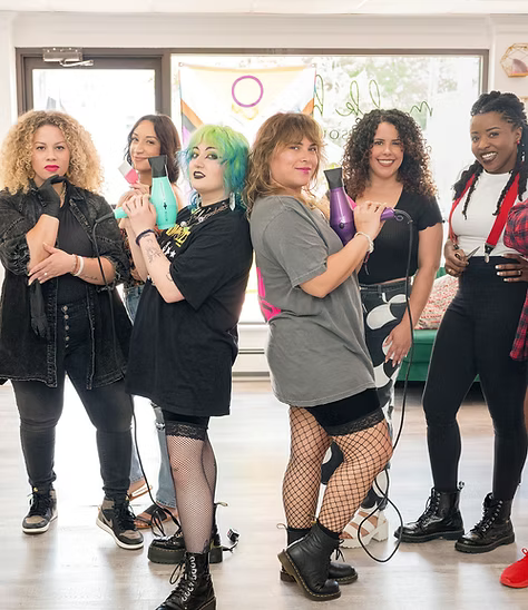 Group of people holding hair dryers, posing indoors. Some have colored hair and unique outfits.