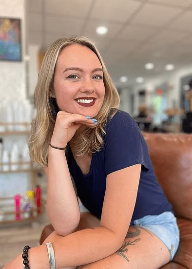 Woman with blonde hair and dark lipstick, smiling and resting chin on hand in a salon.