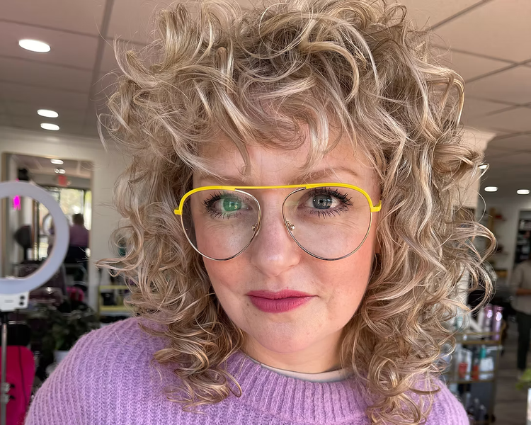 Woman with curly blonde hair wearing yellow-rimmed glasses and a light purple sweater.