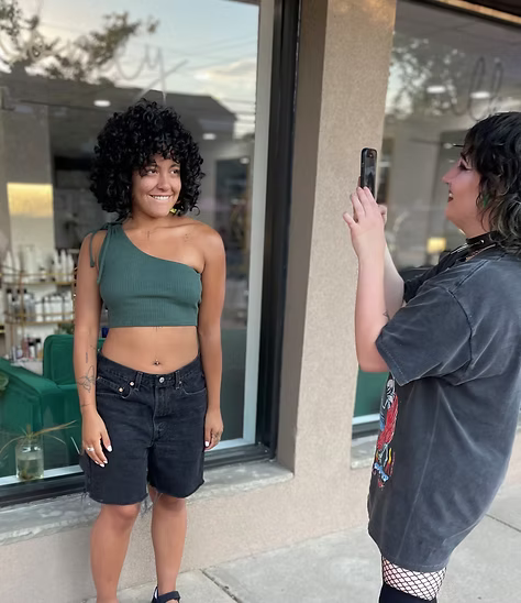 Woman poses in front of a shop while another person takes a photo. The woman wears a green top and shorts.