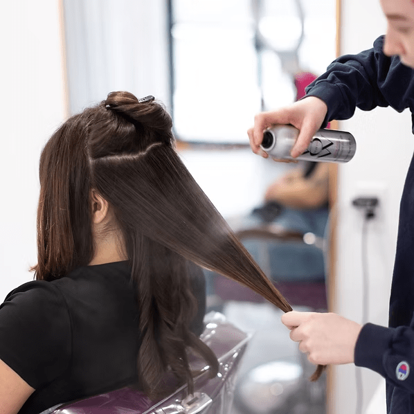 Hairdresser spraying hair product on client's long, dark hair in a salon.