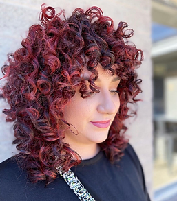 Woman with red curly hair and bangs, wearing a black shirt, leaning against a wall.