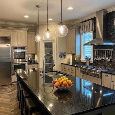 A modern kitchen with a large black island, stainless steel appliances, and globe pendant lights. A bowl of fruit sits on the island's surface.