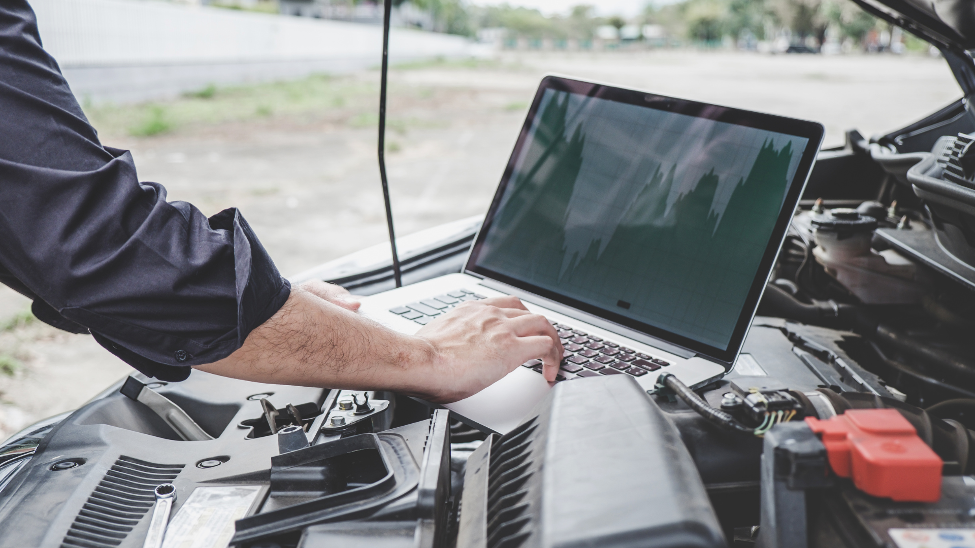 A man is working on a laptop computer under the hood of a car.
