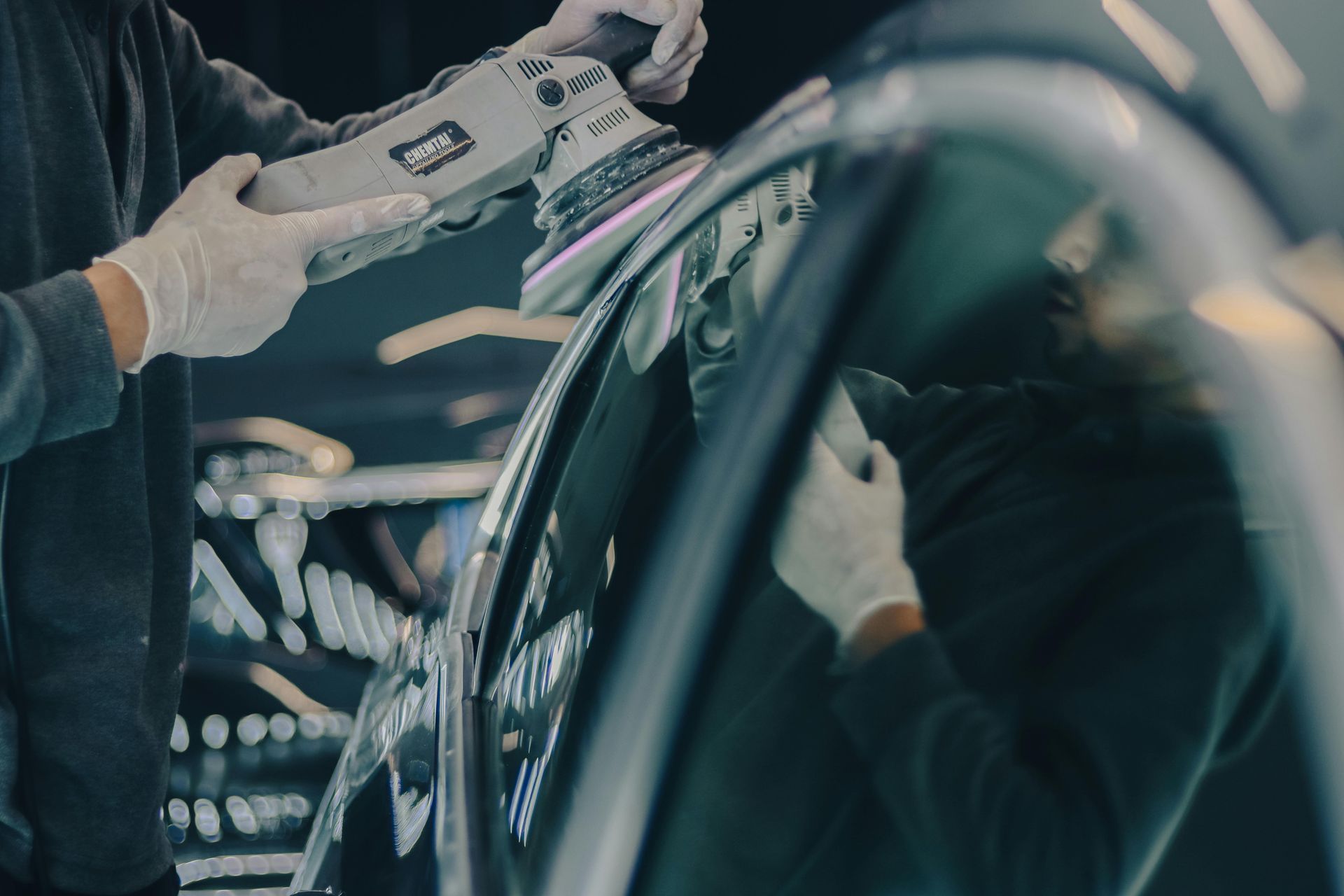 Person polishing car window with a machine, wearing gloves, in a workshop.