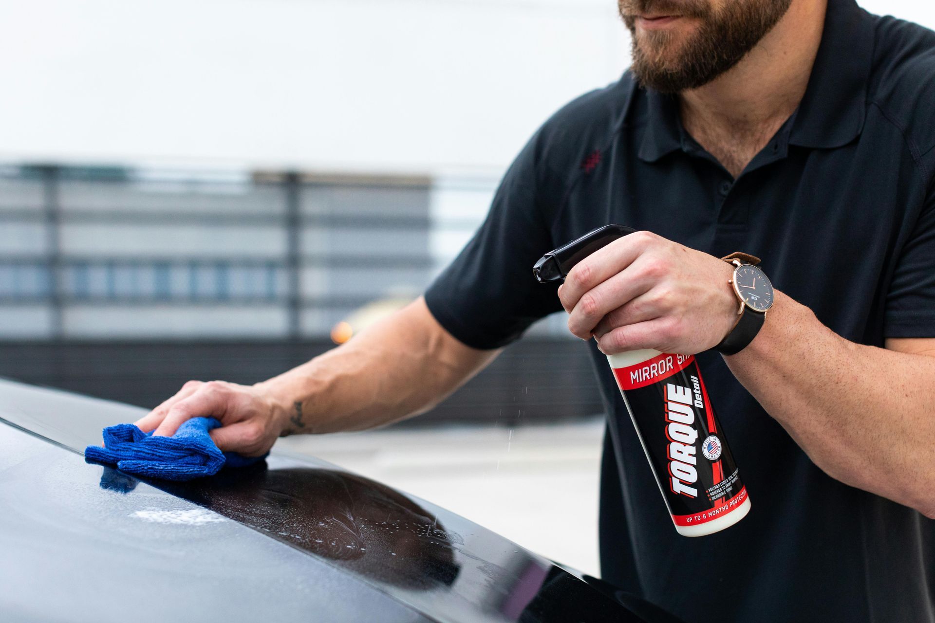Man cleaning a black car with blue cloth and spray bottle labeled