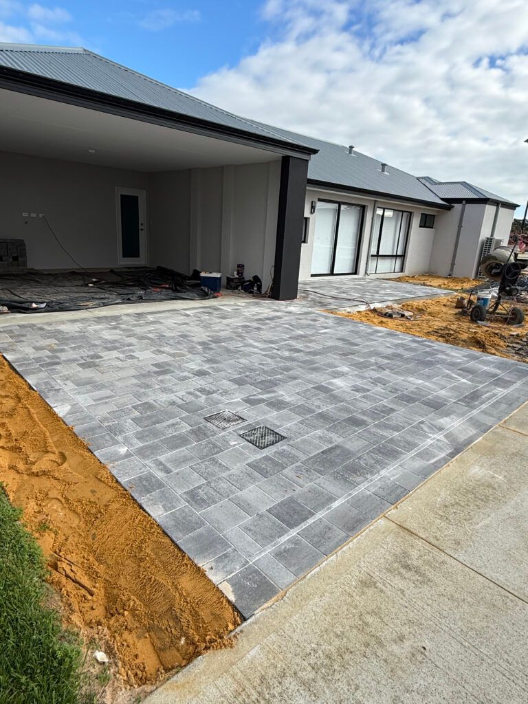 A new grey paver patio under construction outside a modern house with a covered outdoor area and exposed dirt.