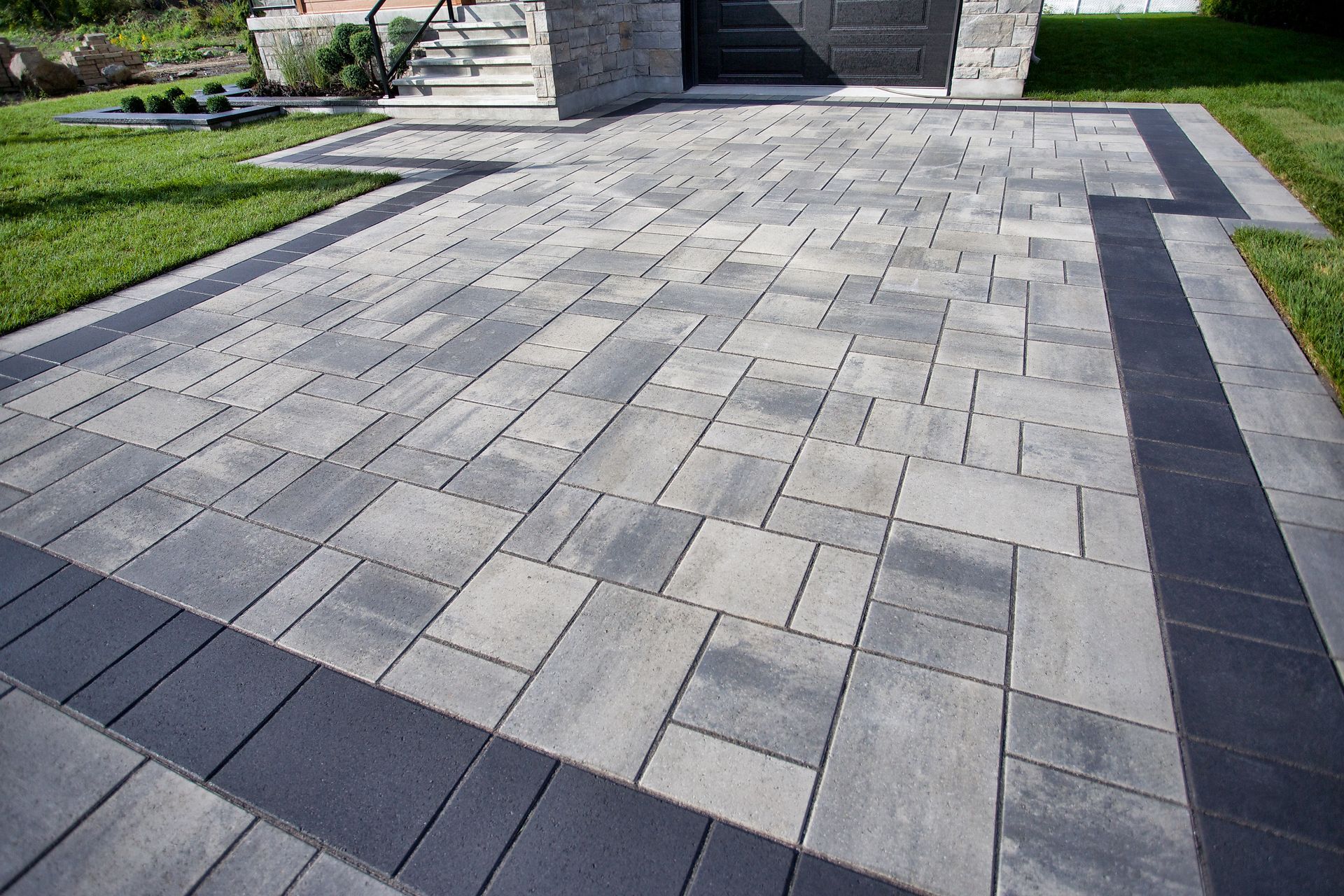 A paved driveway with a mix of grey rectangular pavers framed by a dark charcoal border, leading to a house entrance.