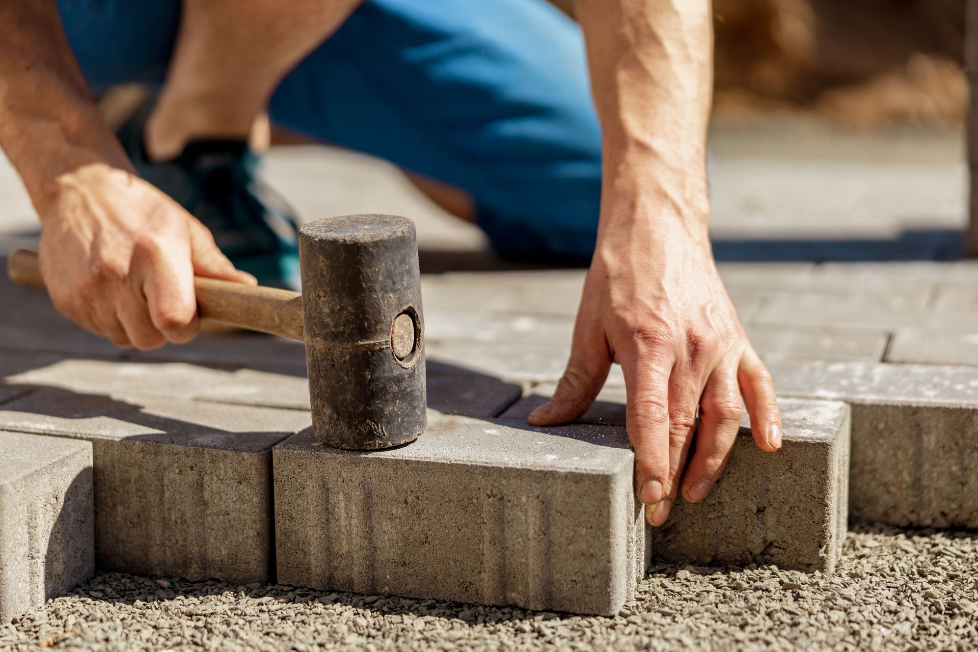A person uses a rubber mallet to secure a gray paving stone into a bed of gravel.