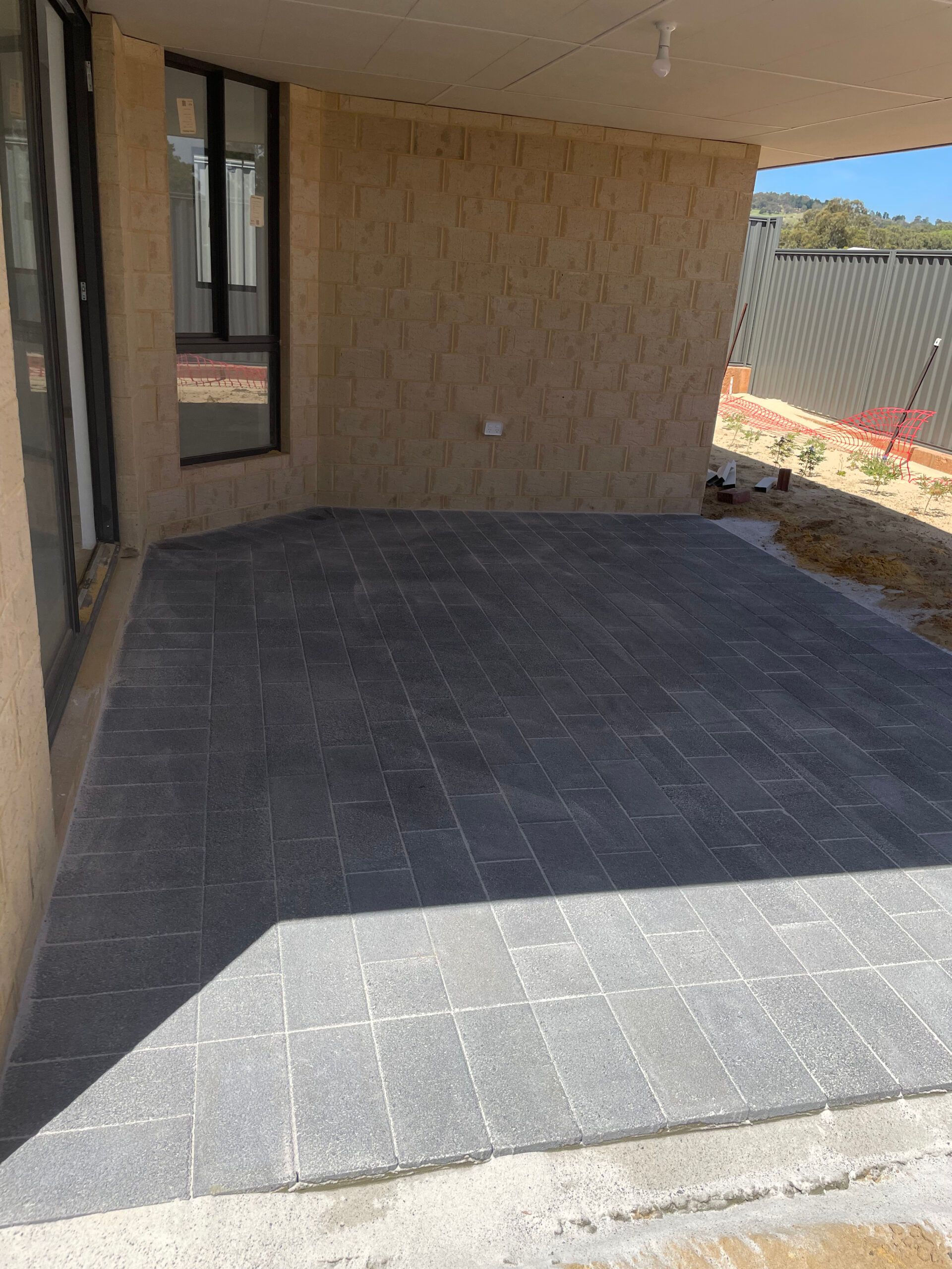 A patio paved with dark gray rectangular pavers under a covered porch area with beige walls and a sliding glass door.