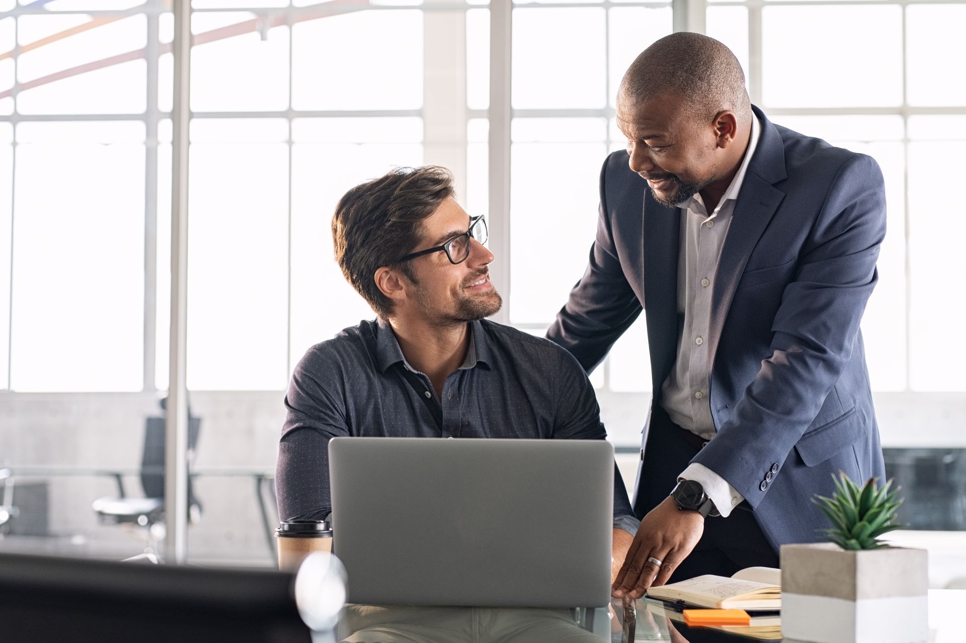 Two men in an office looking at a laptop. One is standing, pointing. Both appear to be smiling. Two men in an office looking at a laptop. One is standing, pointing. Both appear to be smiling.