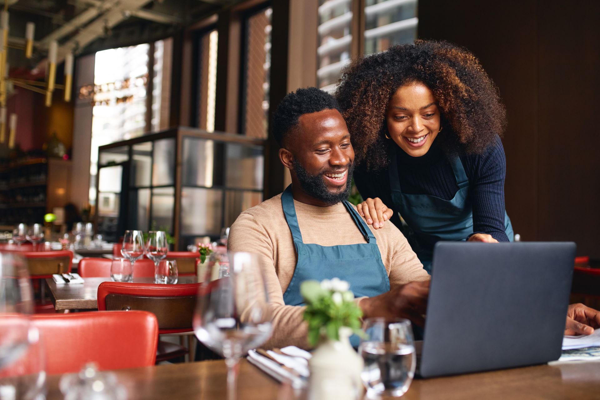 Restaurant owners looking at a laptop together; one is sitting, one standing; in a restaurant setting.