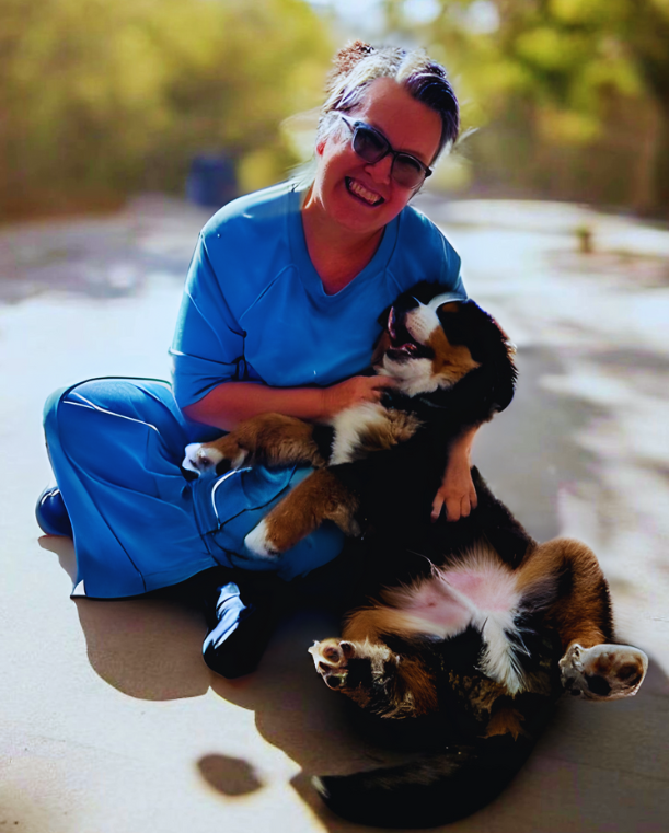 Woman smiles, holding a dog at an office desk with a laptop. Bright room with large windows.