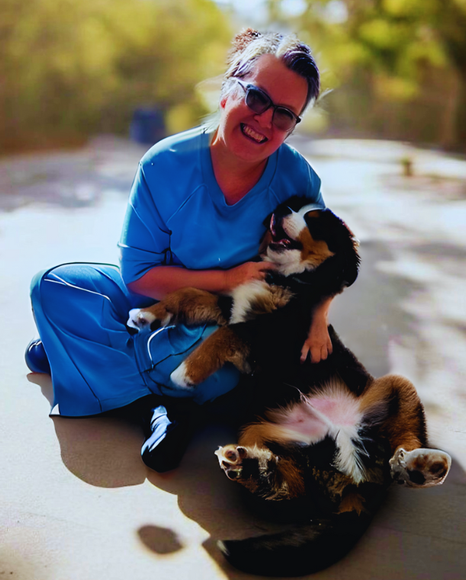 Woman smiles, holding a dog at an office desk with a laptop. Bright room with large windows.