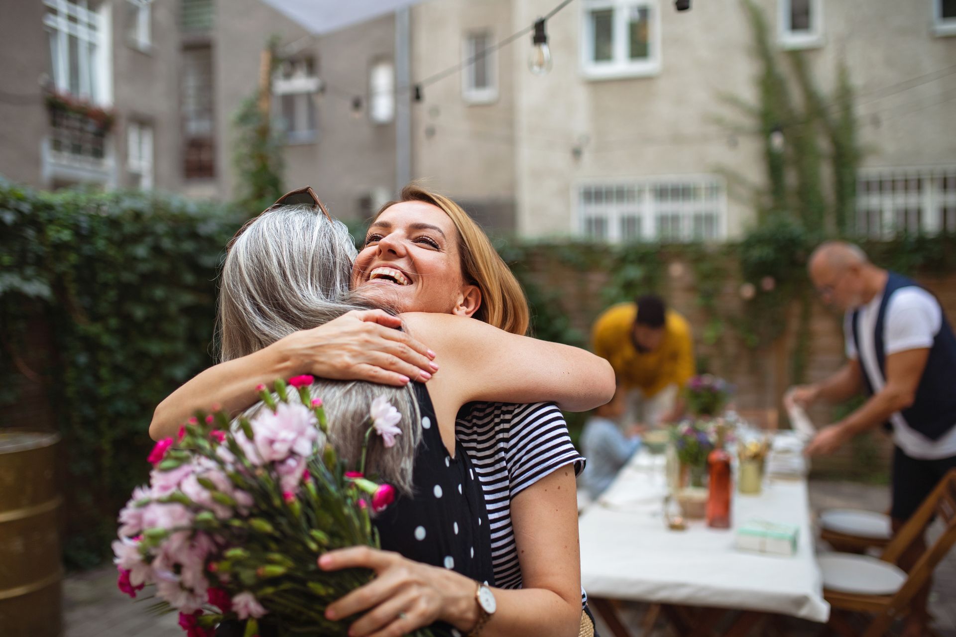 Two women embracing, one holding flowers, smiling widely in a backyard setting.