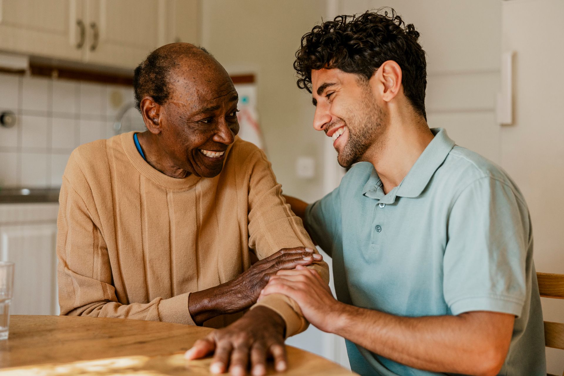 An older Black man and younger man smile, with the younger man's hand on the older man's arm, indoors.