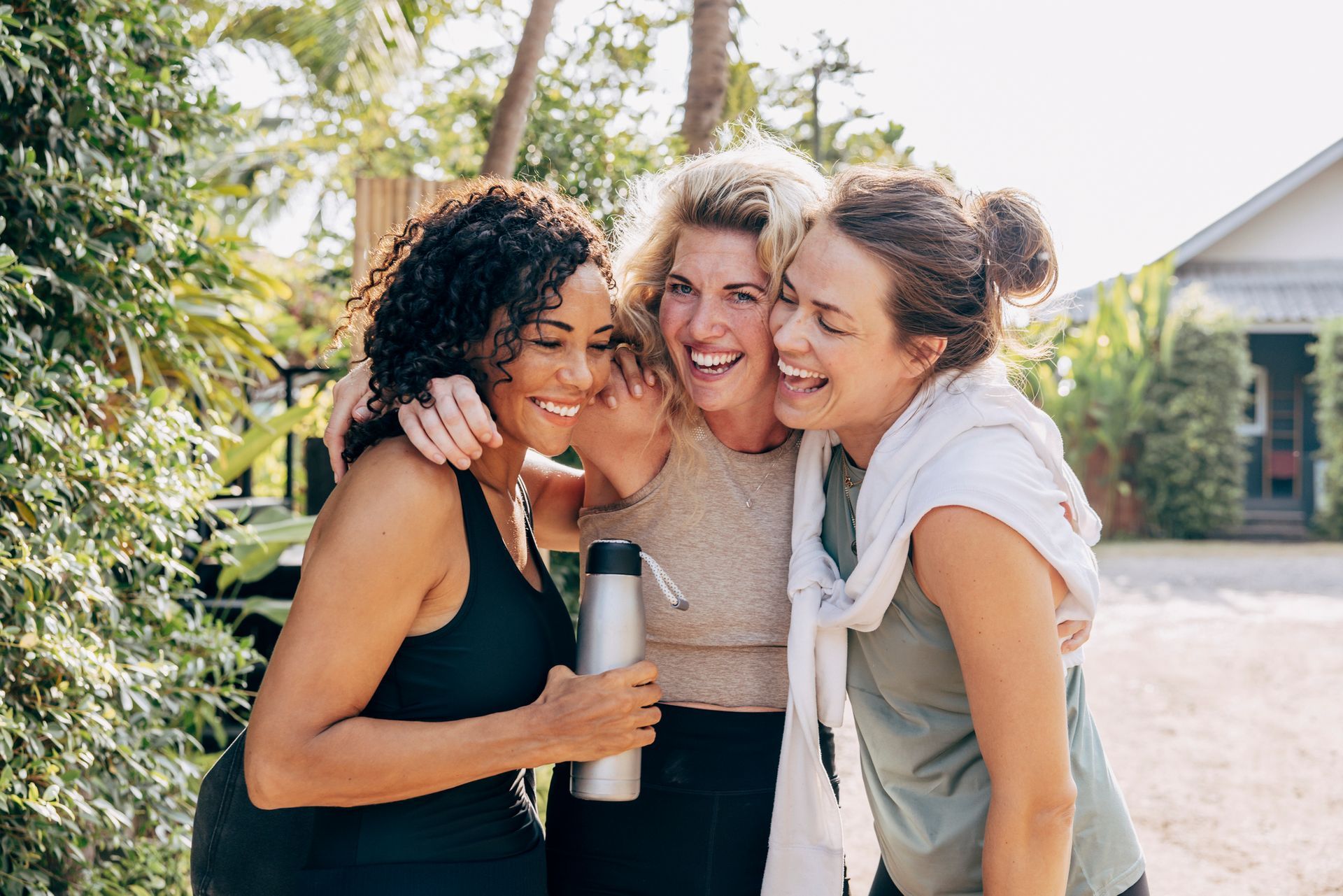 Three women in workout clothes, smiling and hugging outdoors. One holds a water bottle.