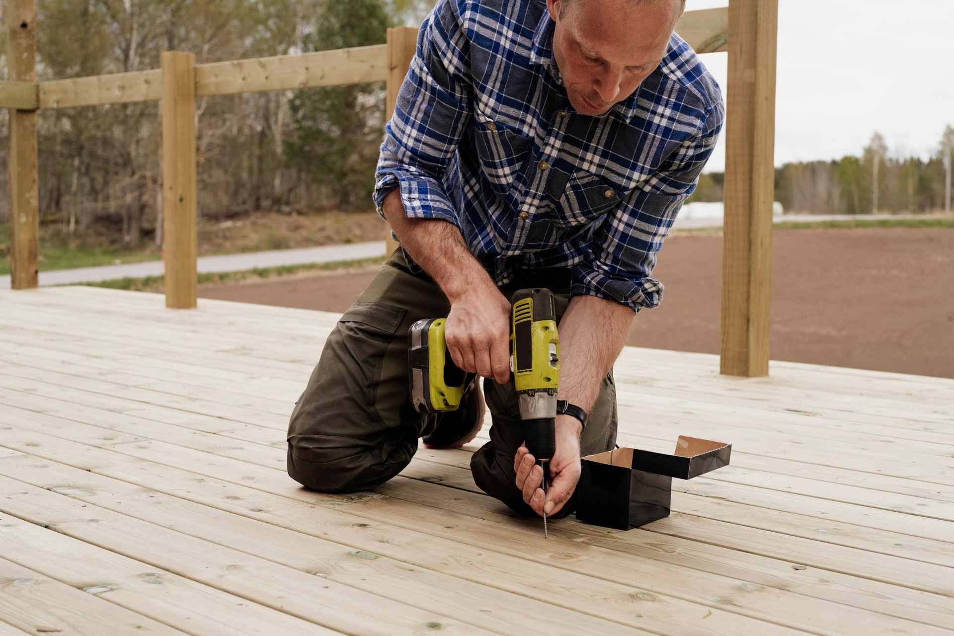 A worker is securing a screw on a wooden deck with a power drill outdoors.