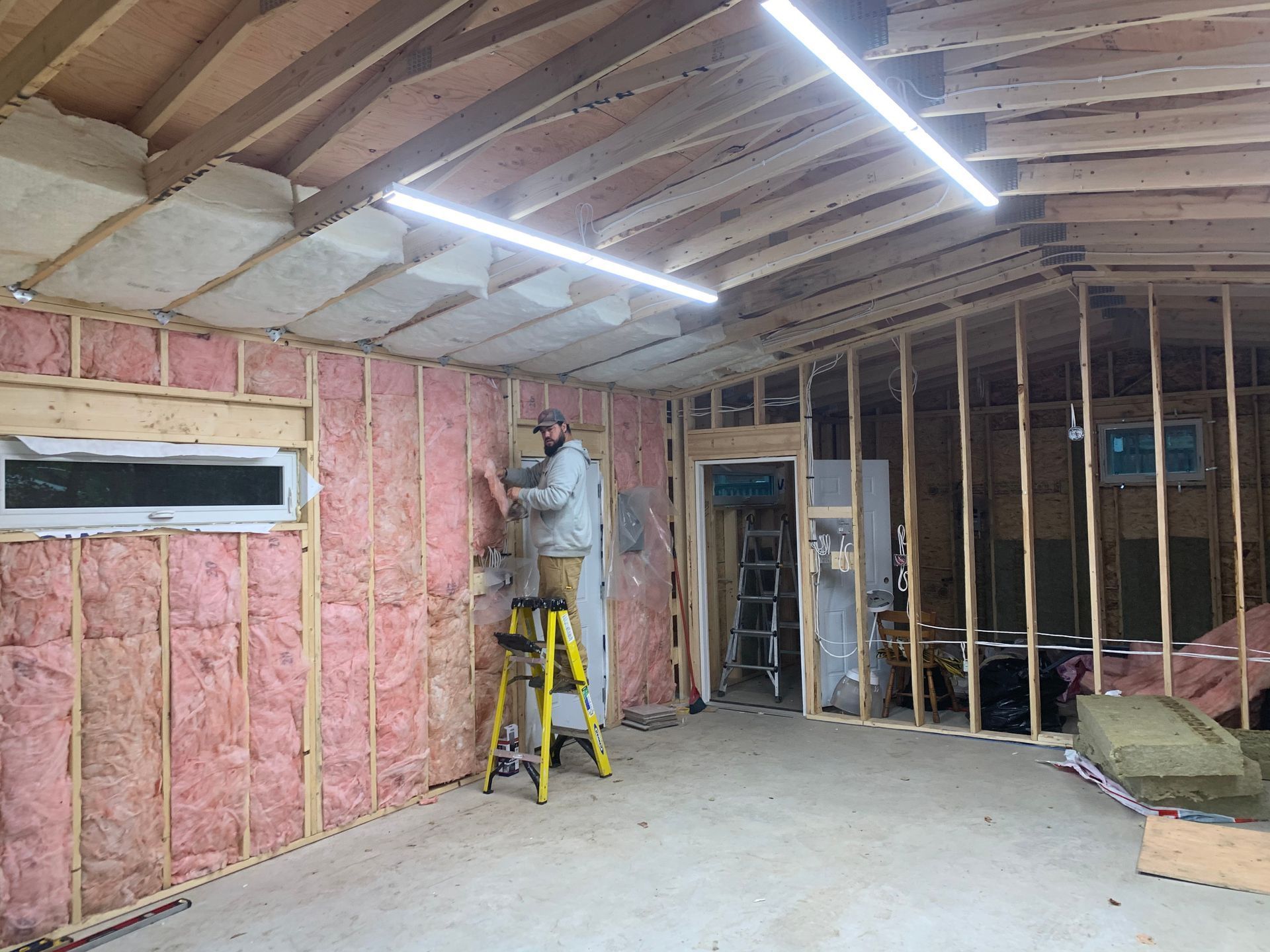 Man on a ladder installing pink insulation in a partially built interior, lit by a fluorescent light.