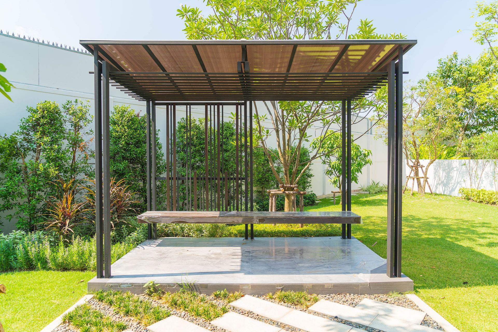 Modern metal gazebo with a bench on a stone patio in a grassy yard.