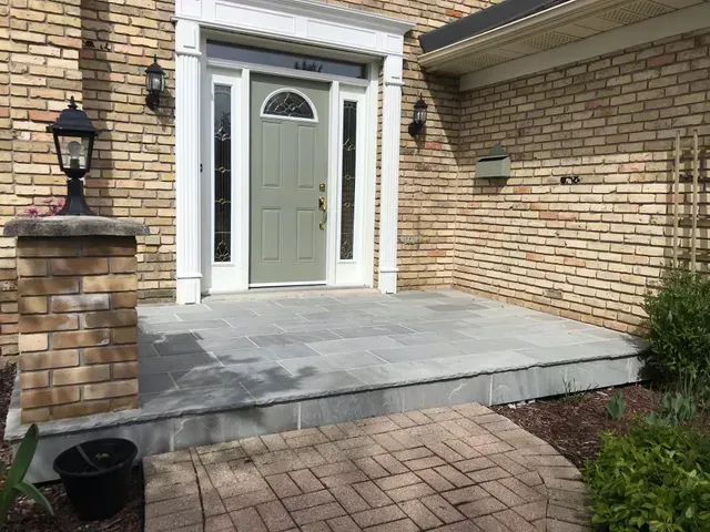 Brick home entrance with gray stone steps and porch. Green door, lamps.