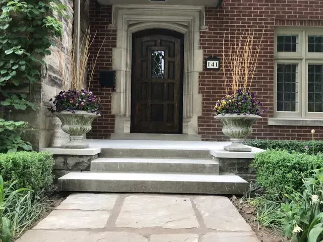 Brick house entrance with stone steps, dark wood door, urn planters, and a walkway.