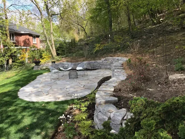 Stone patio with steps and fire pit, surrounded by grass and trees on a hillside.