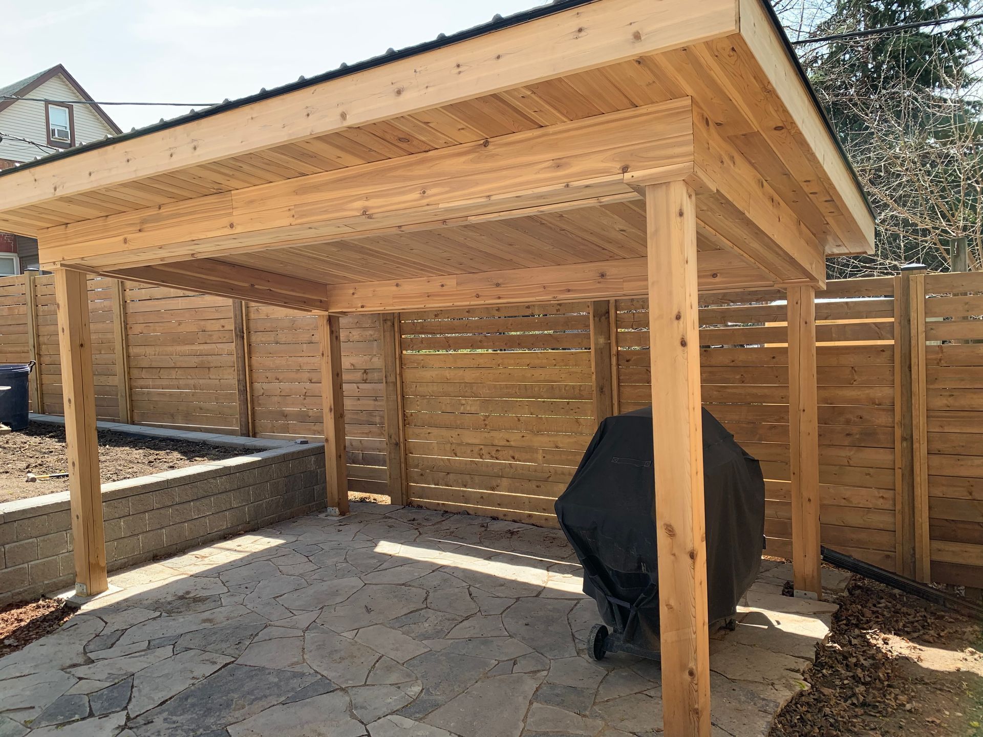 Wooden pergola with a grill on a stone patio, next to a horizontal slat fence.