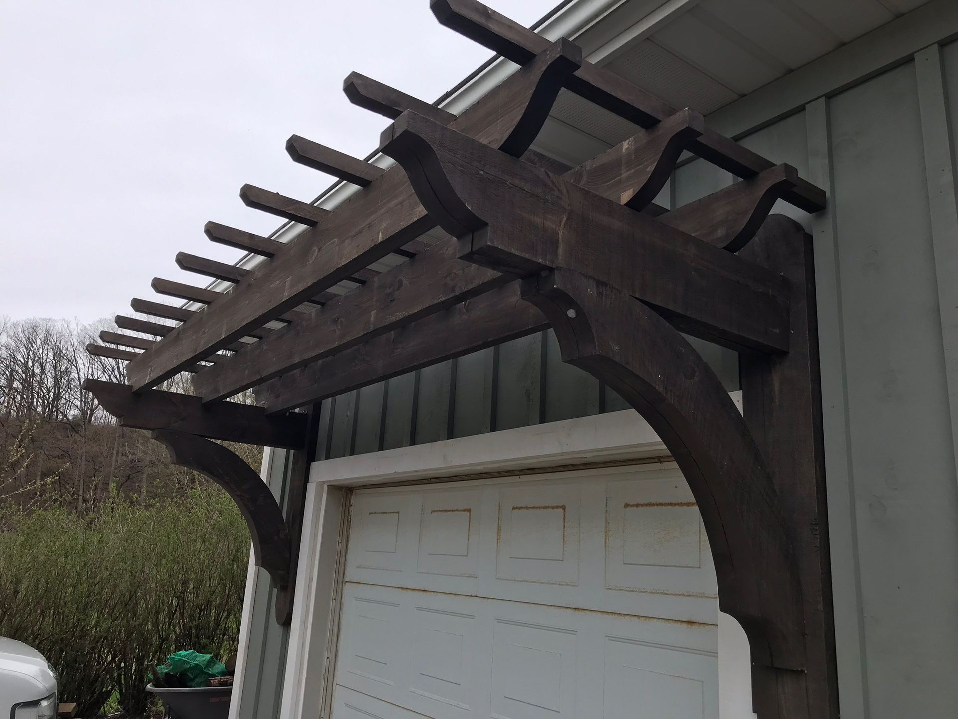 Wooden pergola above a white garage door, attached to a light green building.