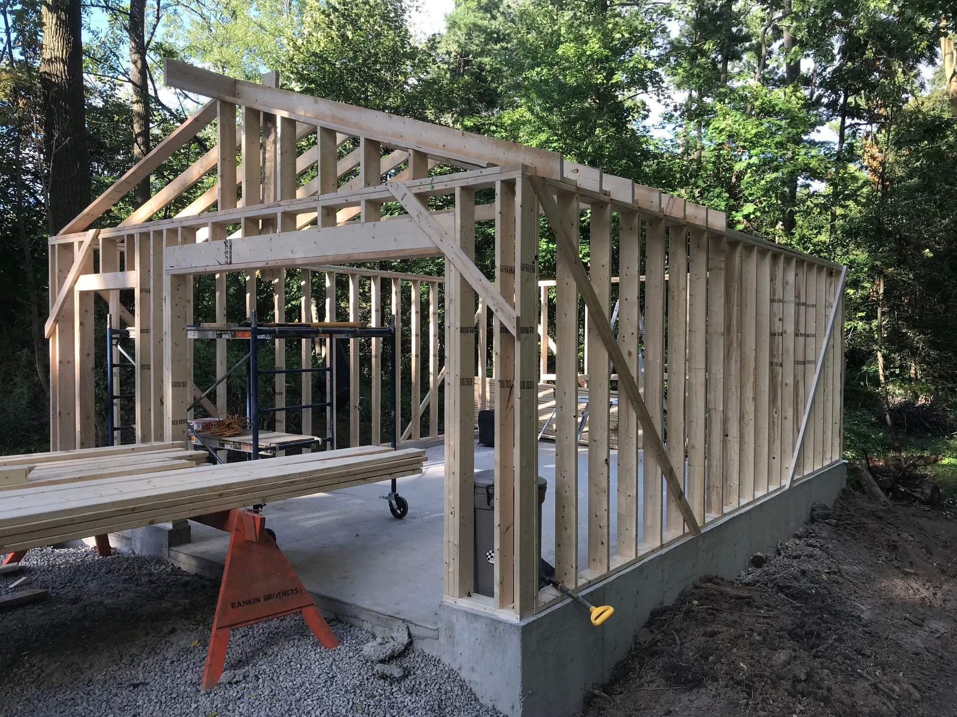 Wooden frame of a building under construction, on a concrete base. Trees in the background.