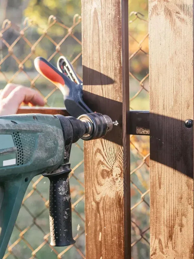 A person using a power drill on a wooden fence post, clamped in place.