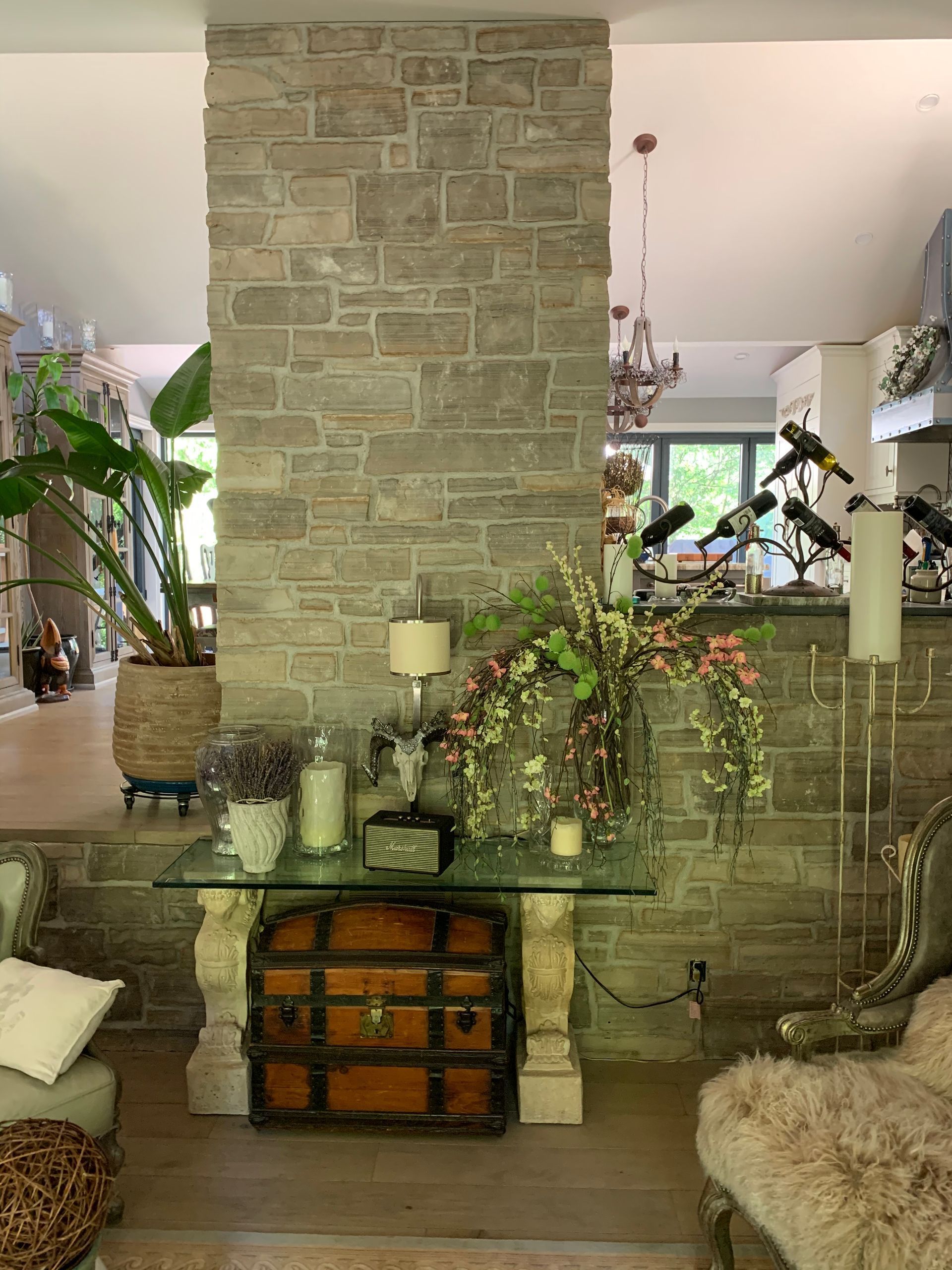 Stone pillar with glass-topped table holding flowers, candles, and a trunk. Living room setting with plants.