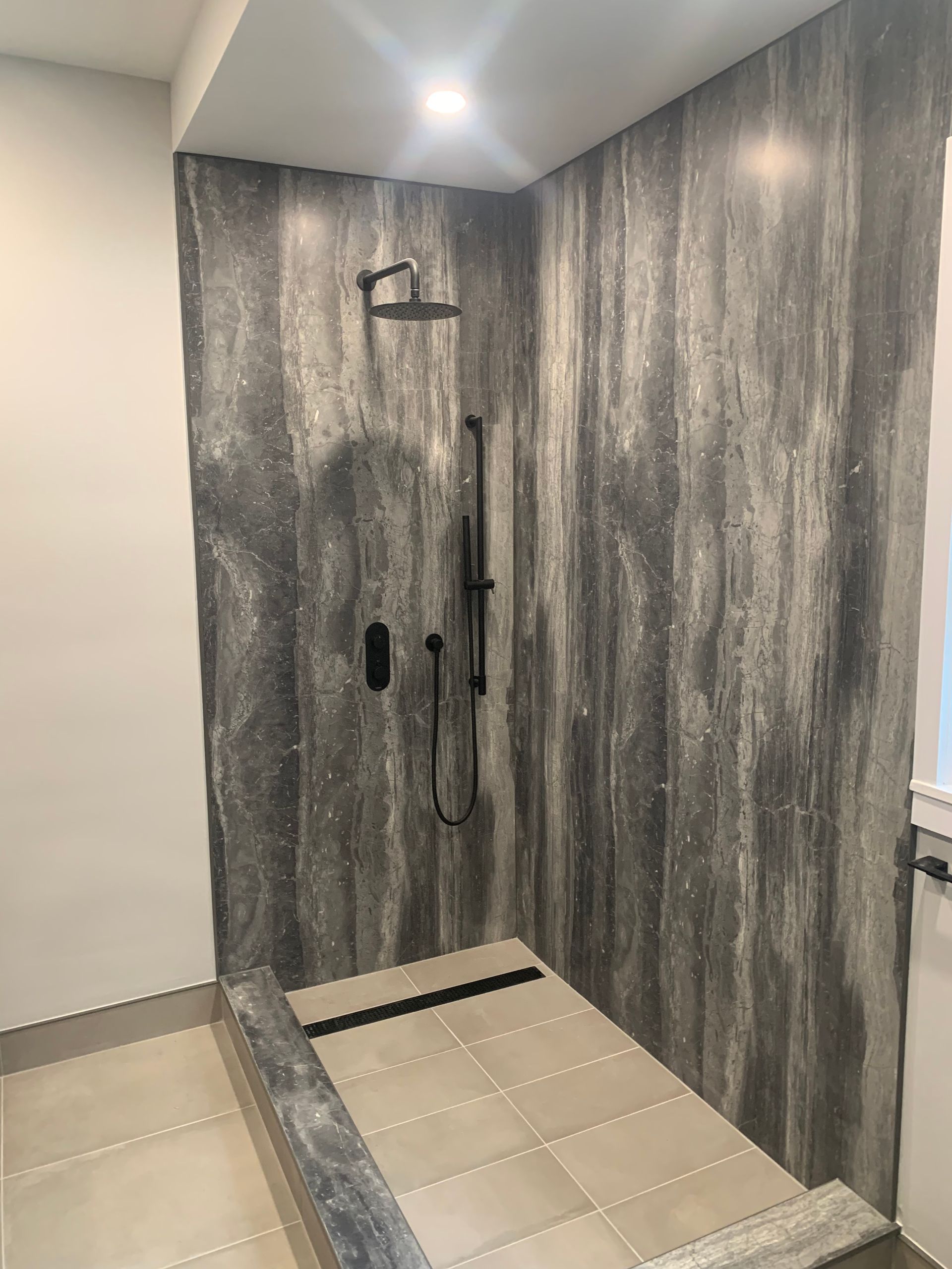Shower stall with textured gray wall panels, black shower fixtures, and light-colored tiled floor.