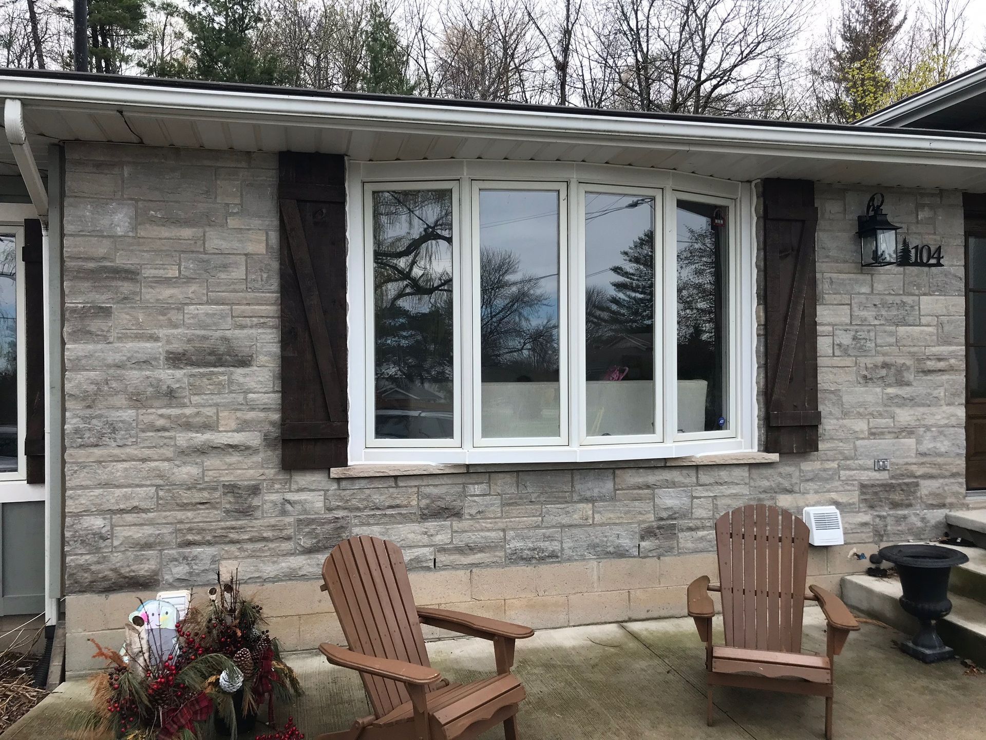 Stone house exterior with white-framed bay window, brown shutters, and two wooden chairs.