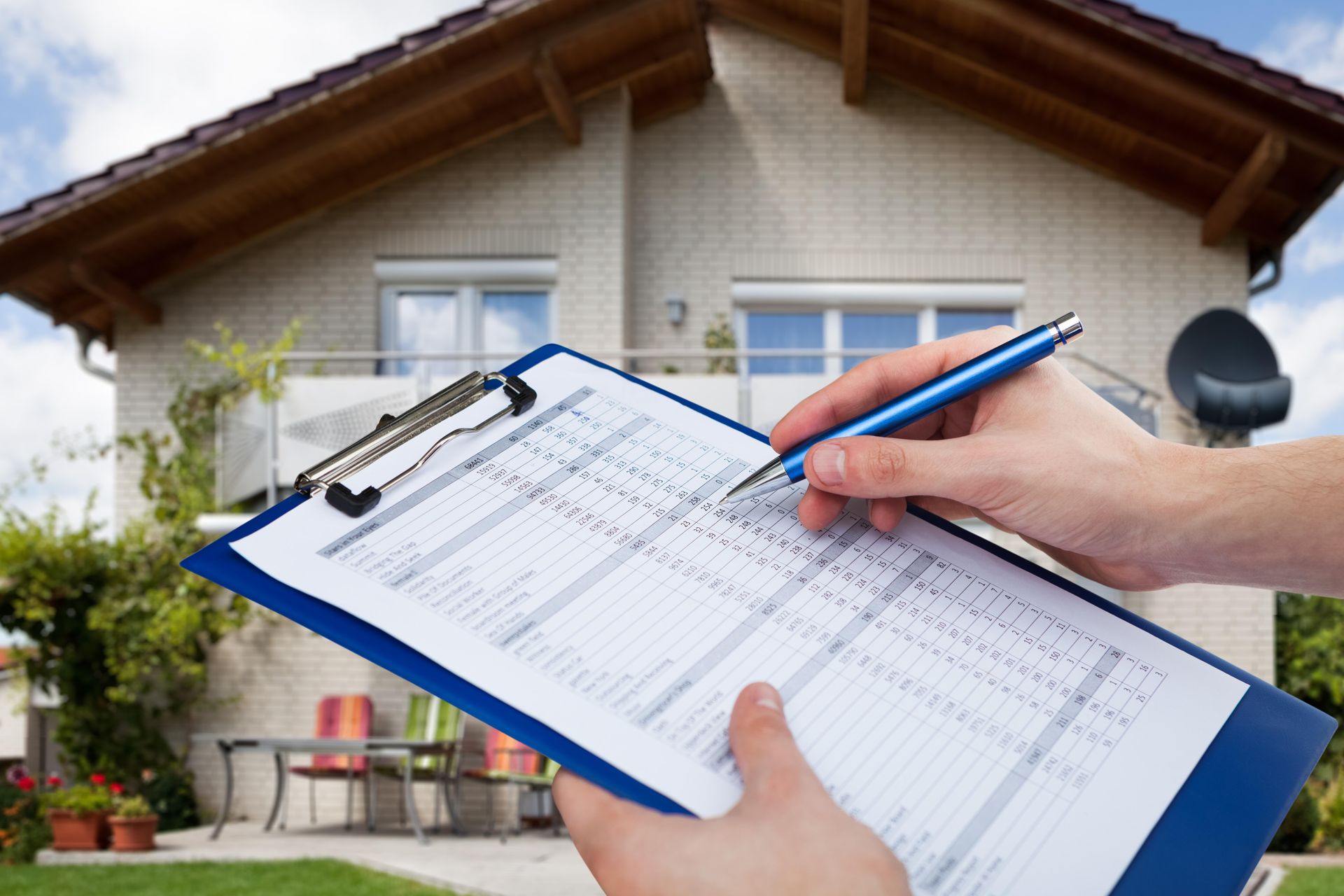 Person holding clipboard and pen, inspecting a house in a green yard, on a sunny day.