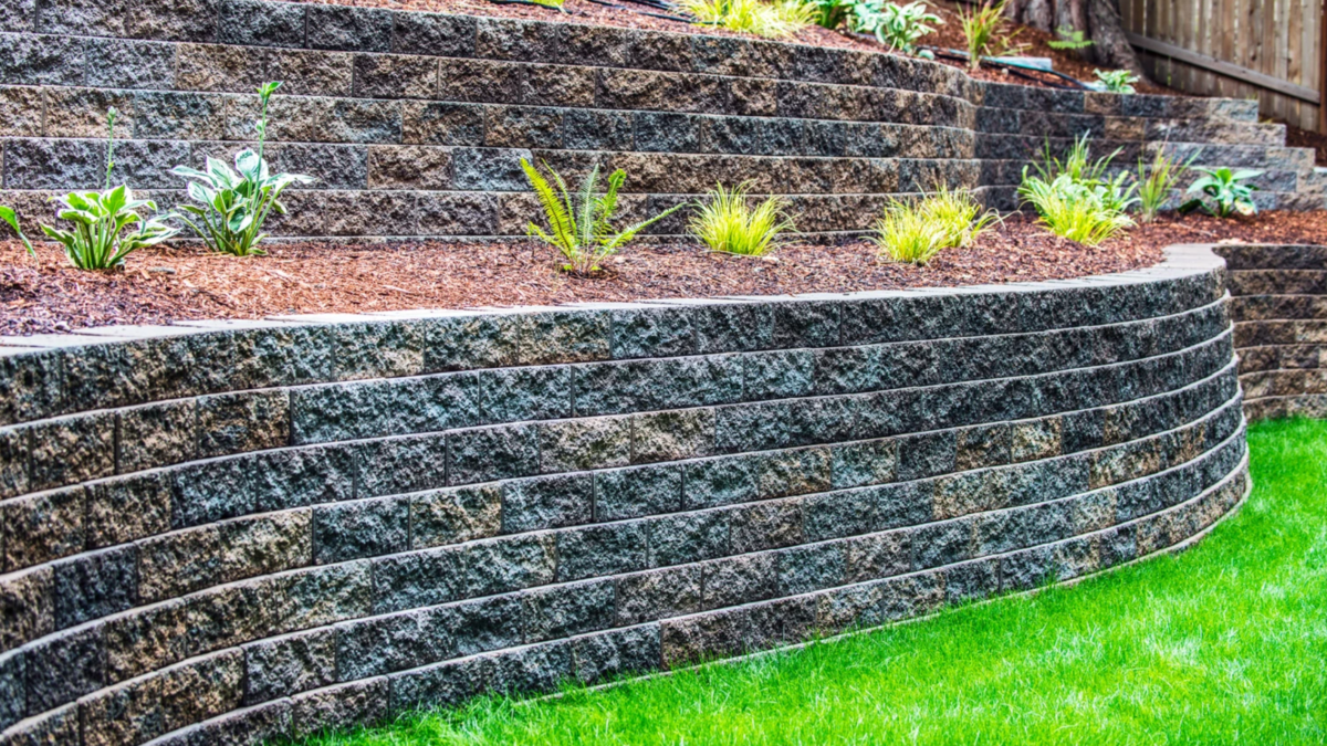 Curved, tiered retaining wall made of dark, textured blocks, with plants and green grass.